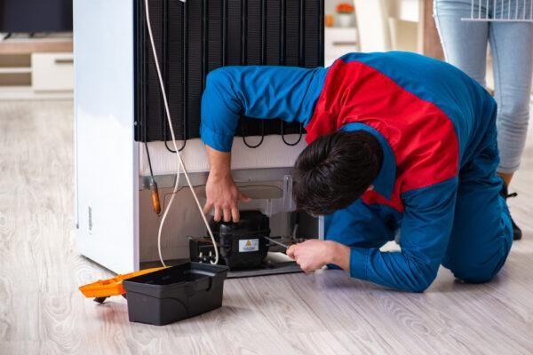 A technician kneels to repair a refrigerator compressor in a Naperville, IL home—highlighting the appliance repair expertise of Unique Repair Services.
